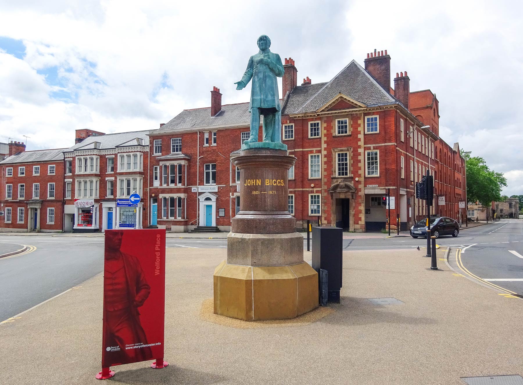 Photograph of the John Biggs statue green tinged, in the middle with one arm slighty out and hand open, on Welford Place a few metres past the end of Lower New Walk